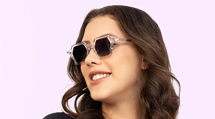 A smiling woman wearing modern hexagonal sunglasses with clear transparent frames and dark lenses, positioned against a soft lavender gradient banner.
