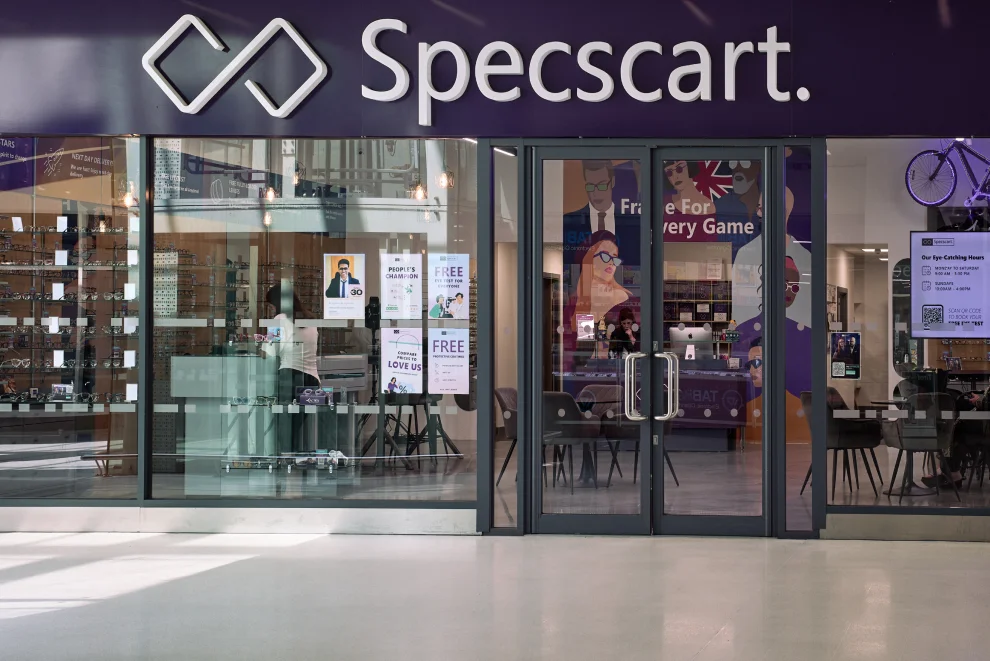 Exterior of Specscart Walkden store featuring a glowing logo, sleek glass entrance, and posters advertising free eye tests in the vibrant interior.