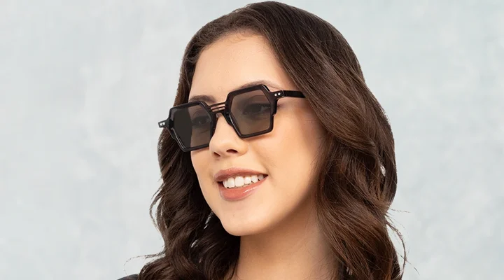 A smiling woman wearing black hexagonal sunglasses with a unique triple-bridge design, posed against a textured light blue and grey banner background.