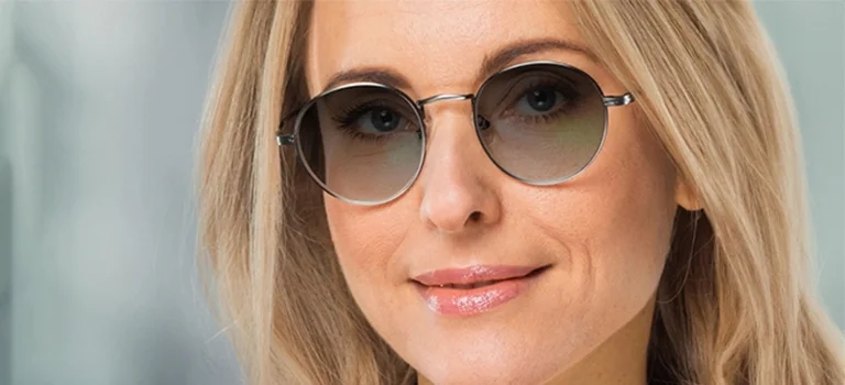 Varifocal Sunglasses A smiling blonde woman wearing elegant round metal sunglasses with gradient varifocal lenses, set against a soft, out-of-focus professional backdrop.